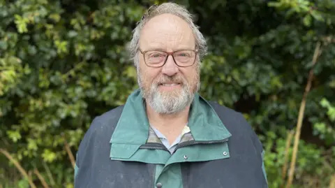 Neil Pullen from Wiltshire Wildlife Trust standing on the edge of Nightingale Wood smiling with a grey beard and brown glasses