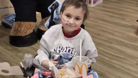 Young girl, 8, sat in a chair with a smile on her face looking straight ahead. She's eating a fast food meal inside a hospital ward.