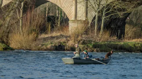 Two men and a dog in a rowing boat, amidst a wide river with a bridge in the background