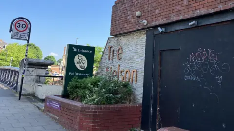 David Freezer/BBC The corner of the Nelson hotel. There is a red and white brick building with a black area with graffiti on it. There is a green sign pointing to the Table Table pub and restaurant. To the left is a 30mph sign on the pavement next to the start of a bridge. 