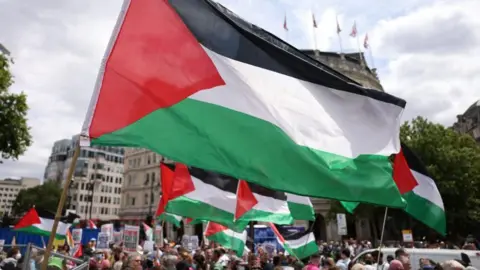 Palestinian flags in green, red, black and white are fluttering against a backdrop of a demonstration in support of Palestine Action in Trafalgar Square, London, in June 2025