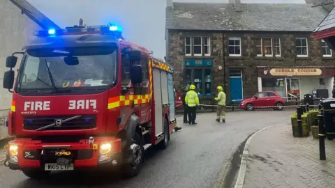 Morva A road closed off with a fire engine at the front and buildings on the road behind.