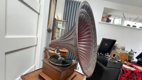 The horn gramophone sits on a little side table. The decorative horn is brown in colour and twists up from a small brown box beneath, with a black record on it.