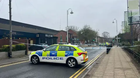 Image shows a police car blocking the road outside the Wall of Fame pub in Formby, Merseyside. There is also police tape across the road.