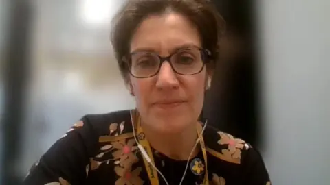 Sarah Holmes look at the camera during a video call. She has short brown hair, glasses and is wearing a black top with a floral print