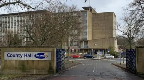 An outside view of Durham County Hall from outside the gates. Cars can be seen parked outside the large grey building.