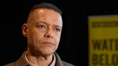 Martin Giles/BBC Cliver Lewis is left of the shot, standing inside and wearing a checked shirt, jumper and tweed jacket. He has short dark hair and there is a yellow sign in the background.