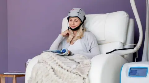 A woman sat in a chair wearing a cooling cap. She is reading a magazine and has a blanket over her. 