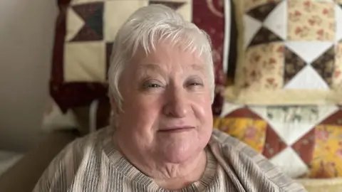Margaret Galloway has grey hair parted slightly to the side. Quilted, patterned cushions are visible behind her.