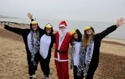 St Elizabeth Hospice Four people dressed in penguin outfits on Felixstowe beach, with a person in a Father Christmas outfit in the middle