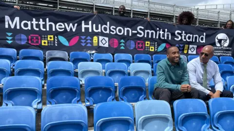 Hudson-Smith and his dad are sitting on two of the blue seats on the right. The faces of three people are visible at the top of the image behind the banner with the words Matthew Hudson-Smith Stand.