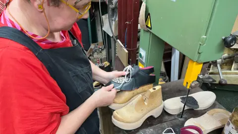 A wooden last is being covered by a piece of black and white leather with decorative laces in, next to it is the wooden sole of a shoe 