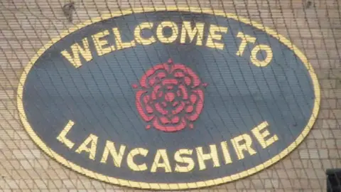 M J Richardson/Geograph Oval sign saying "welcome to Lancashire" with a red rose in the middle. the writing is gold on a black background