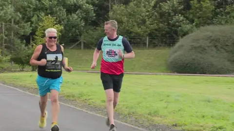 Debbie running in a park wearing a vest that reads "Stop Glioblastoma". Graeme is running wearing a vest that reads "The Brain Tumour Charity".