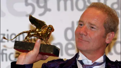 Getty Images Peter Mullan, clean shaven this time, holds the Golden Lion trophy up and looks at it. He is wearing a dark purple kilt jacket with white shirt and mauve cravat.