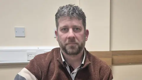 BBC Donal 'Mandy' Kelly has short black hair and is wearing a multi-coloured fleece. He is standing in a beige room with a number of light switches and sockets behind him. 