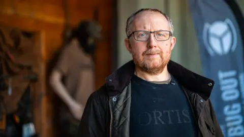 BBC A man with a ginger beard and grey hair stands in the porch of his workshop wearing a dark green jacket over a black shirt. There is a mannequin wearing a gas mask and green army clothes behind him. Rucksacks hang on the wall. 