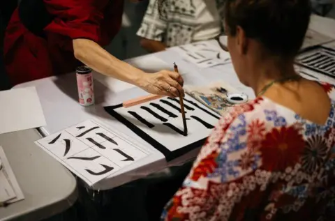 Hyper Japan A woman in a blue, white, and red flowered top learns how to write Japanese characters.