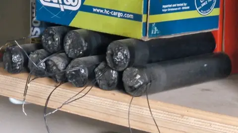 West Yorkshire Police A wooden shelf holding a group of cylindrical objects that appear to be tightly wrapped in dark material. Each cylinder has exposed wiring extending from one end, and the ends appear sealed with a rough, compressed-looking substance. The cylinders are stacked in two layers, with several placed side by side and others positioned on top. A cardboard box with blue, green, and yellow branding sits directly above some of the cylinders.