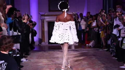 Getty Images A model wearing one of Shonagh's designs at London Fashion Week. The off-the-shoulder mini dress, pictured from the back, is made from white yarn, has long flowing sleeves. The model wears a matching crown-style headband and walks down the catwalk being photographed by audience members on their smartphones.