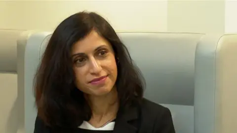 A woman with thick black hair, dark pink lipstick and a black blazer is looking towards the right hand side of the camera. She sits on a light beige chair.