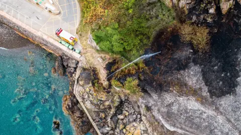 States of Jersey Fire and Rescue Service Aerial view of wildfire at Bonne Nuit in 2018. A fire engine is parked in a car park next to the open land which had been damaged by the flames. Firefighters are standing on the edge of the land, next to the water, using a hose reel jet to spray water over the area. 