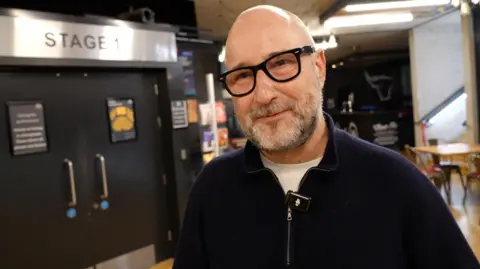 A man stands inside a modern theatre lobby. He is wearing a dark zip‑up sweater. Behind him there are large double doors marked “Stage 1”. Posters and notices are displayed on and around the doors. To the right, the lobby opens into a seating area with wooden tables and chairs, lit by warm overhead lighting. 
