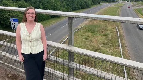 Amy Holmes/BBC A woman standing by a railing on a footbridge above a dual carriageway, with trees, road signs, and passing cars under an overcast sky.
