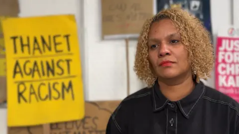 Jadzia Samuel/BBC A woman stands in front of placards which reads Thanet against racism