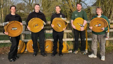 Dunstable Community Fire Station A group of five people, all holding yellow fire hoses, standing by a wooden fence. Three are wearing black trousers, boots and black tops. The other two have green jumpers on. A man, second from the right, has black jeans on, and a woman next to him has leopard print trousers. They are all smiling. Trees are behind them. 
