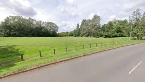 Google A general view of a section of Ladybridge Park. It shows a large grassed area surrounded by trees. A single carriageway road runs next to it.