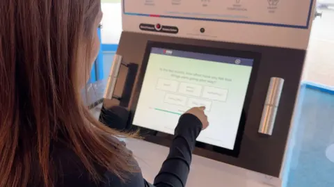 You can see the side of an anonymous woman's face with long dark hair sitting in front of a machine with a screen, which she is tapping with her finger. 