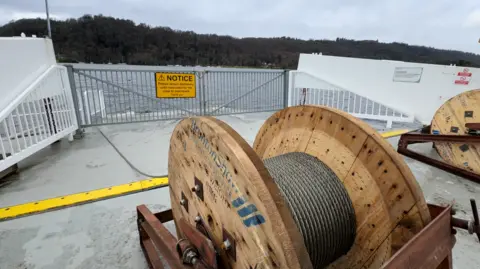 In the foreground there's a large wooden reel, with a thick grey cable wrapped around it. The cable runs across the grey deck, under a gate with a yellow warning notice on it, and into the lake. In the background there's a lake and a forest on the far bank.