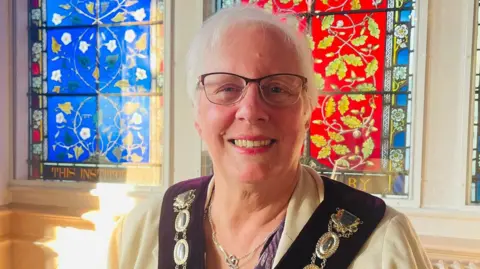 Gordon King A woman - Carole King - with short grey hair and glasses looking at the camera and smiling - wearing a mayoral civic badge and standing in front of red and blue stained-glass windows. 