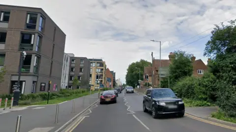 Sturry Road in Canterbury, with mid-rise buildings on one side and some houses and trees on the other. There are queues of traffic in both directions.