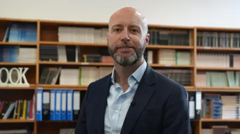 BBC/Brijesh Patel Tony Oulton looks astatine  the camera smiling wearing a achromatic  suit   overgarment   and airy  bluish  shirt. He has a grey and achromatic  abbreviated  beard. Behind him determination   are shelves with rows of coloured books and folder. 