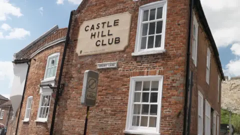 The Castle Hill Club is a tall red bricked building, it has a large beige sign with black bold writing with its name on. It has white sash windows.