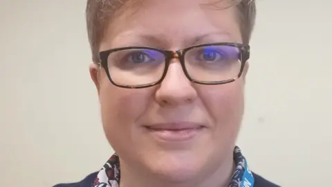 Kettering General Hospital A woman with short blonde hair, glasses and wearing a shirt and jumper looks at the camera