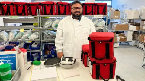 Syed Mushtaq standing in the lab and smiling at the camera. He is wearing a white coat and is surrounded by the red cool boxes his team uses to bring eyes to the lab for transplant.
