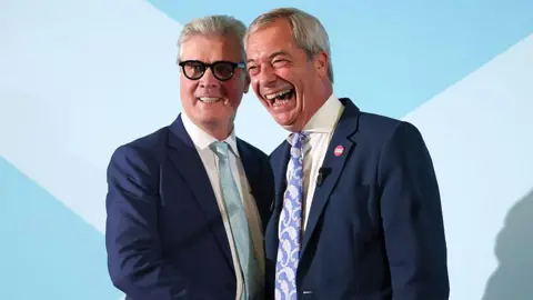 Getty Images Malcolm Offord and Nigel Farage standing close together on a stage. They both have short, grey hair and are wearing blue suits and cream or white shirts. Offord has dark-rimmed glasses and is wearing a light blue tie. He is smiling while Farage, who is laughing, is wearing a paisley-patterned blue and white tie. 