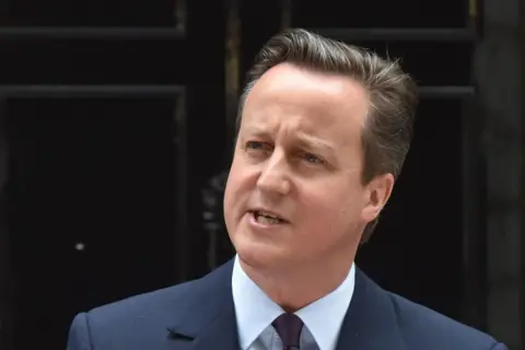 A man in a dark suit, light shirt, and tie stands at a microphone in front of the black doorway of 10 Downing Street. The image is a close-up portrait with the entrance forming a dark background.
