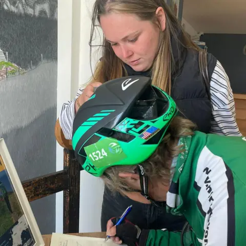 A picture of Peggy and her granddaughter signing the book at Land's End. Peggy is wearing a green helmet and a green jacket. Her granddaughter has long blonde hair and is wearing a striped top and gilet.