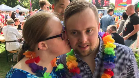 Michael Gallagher stands next to his girlfriend Alison Williams at a Pride event. They are both wearing colourful flower garlands around their necks and Alison kisses Michael's cheek. There are people behind them sitting on tables on grass and Pride banners.