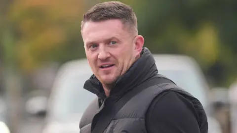 PA Media Political activist Tommy Robinson outside Folkestone Police Station. Robinson is wearing a black jumper and a black gillet. He is looking slightly away from the camera taking the image, has stubble and his hair gelled to one side.