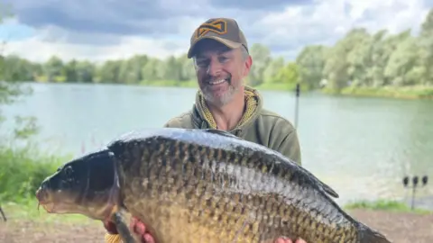 Alyson Noakes Luke Robbins, wearing a dark green hoodie and cap, holds up a large fish to the camera while sat beside a lake.