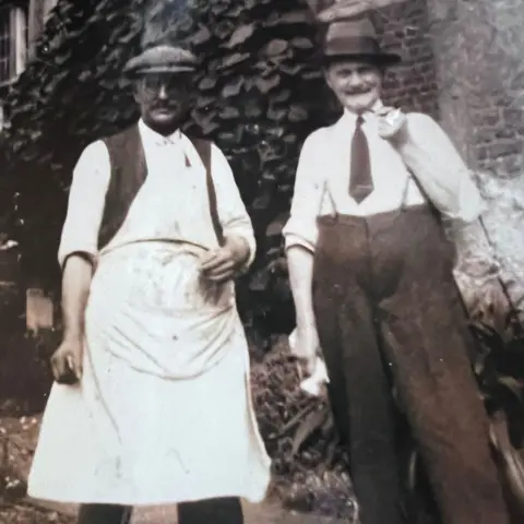 Steve Rowling A black and white picture of two men staring at the camera, the man on the left is wearing a white apron, waistcoat and flat cap, and his companion is wearing a tie, smart trousers, braces and a shirt 