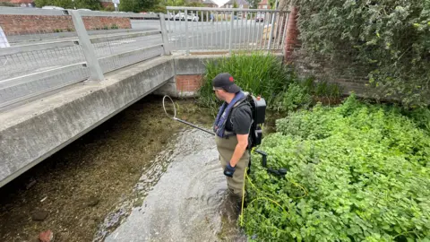 Oliver Southgate from East Yorkshire River Trust. He is standing in a stream and is carrying fishing equipment. He has a backpack on his back.