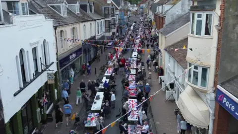 A drone shot of a previous VE Day celebration with tables lined up the street with union jack flags on the tables and hundreds of people milling about.