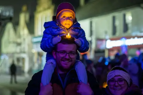 Paul Campbell A young family out in a street in Dingwall in the dark. A child, wrapped up in cosy clothes, is on her dad's shoulders. She has placed a battery-operated candle on her dad's head and the "flame" is lighting up her face.