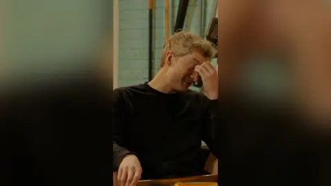 A man with short blond pinches the bridge of his nose as he looks away from a plate of food